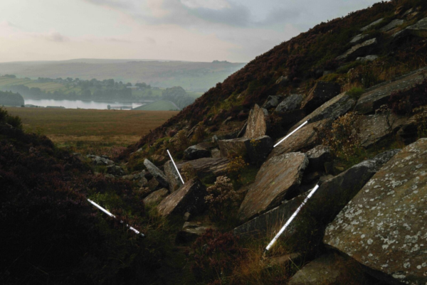 A hillside scattered with large rocks is intersected by thin illuminated light strips, creating a striking contrast against the natural landscape and moody skies.