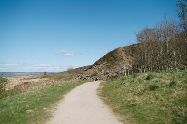 Gravel walking path winding through grassy moorland beside a rocky hillside under a clear blue sky with scattered clouds.