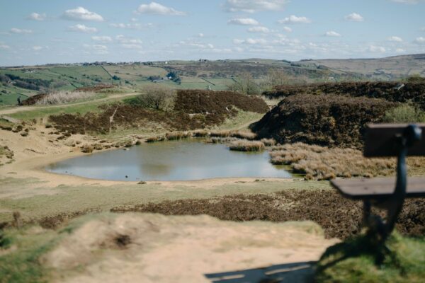 A scenic view over a small pond surrounded by reeds and heather-covered hills, with a wooden bench in the foreground and rolling hills in the distance under a blue sky with scattered clouds.
