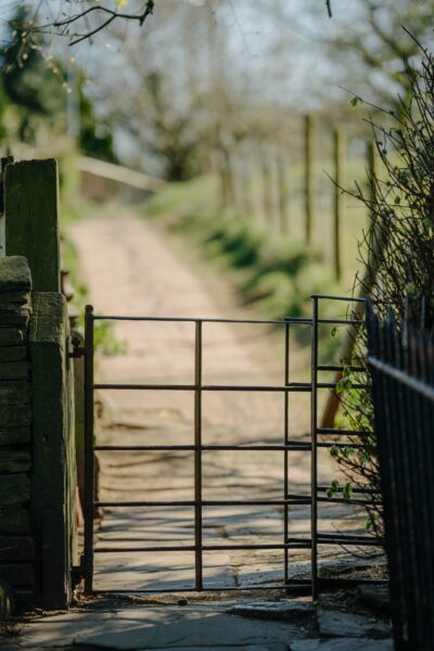 Zoomed-in view of a metal farm gate set between stone walls and a wire fence, opening onto a sunlit rural path flanked by grass and trees.