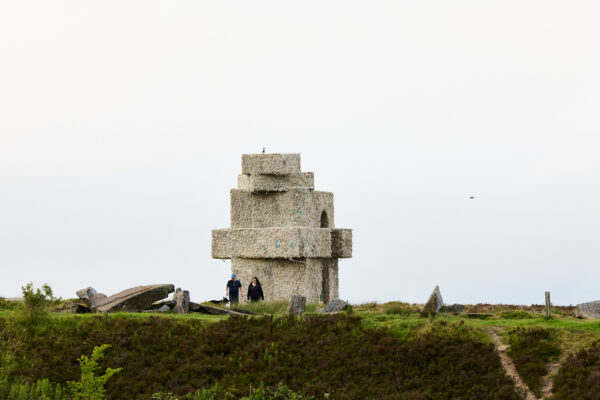 Large stacked oblong shapes forming an arch sculpture made from wool, standing on open moorland beside large scattered stones, with two people walking nearby under an overcast sky.