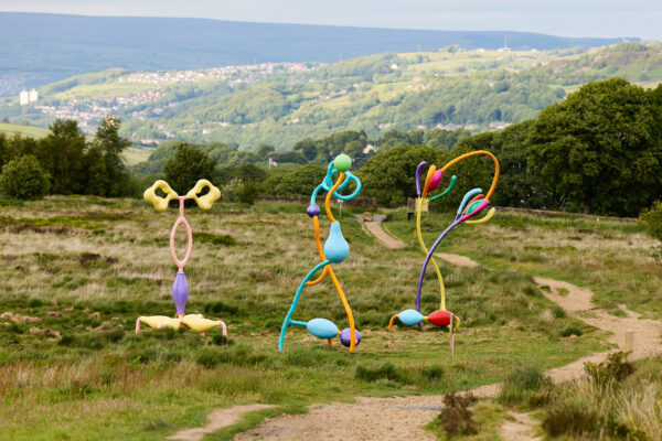 Three abstract sculptures in bright pastel colours stand on a grassy hillside, their looping and organic forms resembling playful plant structures, set against a backdrop of distant hills and woodland under an overcast sky.