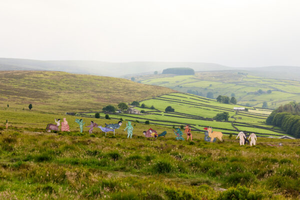 Panoramic view of a grassy landscape dotted with colorful sculptures inspired by medieval animal illustrations, featuring fantastical hybrid creatures stretching across the frame, with stone walls and farmland in the distance.