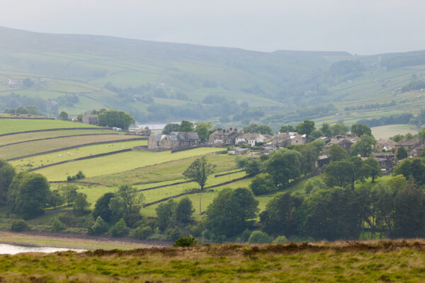 Stone-built village nestled in rolling green fields divided by dry stone walls, with scattered trees and a reservoir visible in the background beneath overcast skies.