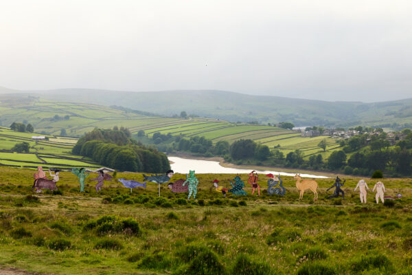 Long line of animal sculptures arranged on grassy hills overlooking a winding reservoir and patchwork fields, featuring fantastical hybrid creatures set against a misty rural landscape.