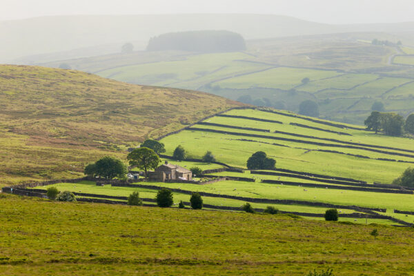 A view from Penistone Hill with misty hills and green farmland in the background. There is an old farmhouse building, made of sand-coloured bricks with flat green space around it for sheep to graze.