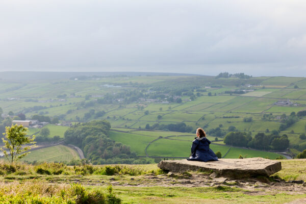 Person in a dark blue coat seated on a large flat rock slab, overlooking green patchwork fields and hedgerows stretching across rolling hills under an overcast sky.