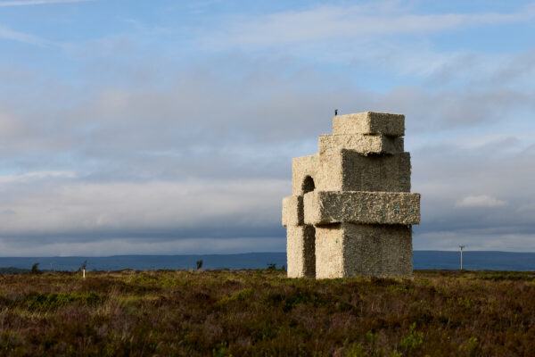 Large stacked oblong shapes, making an arch sculpture made from wool, standing alone on open moorland under a partly cloudy sky with hills in the distance.