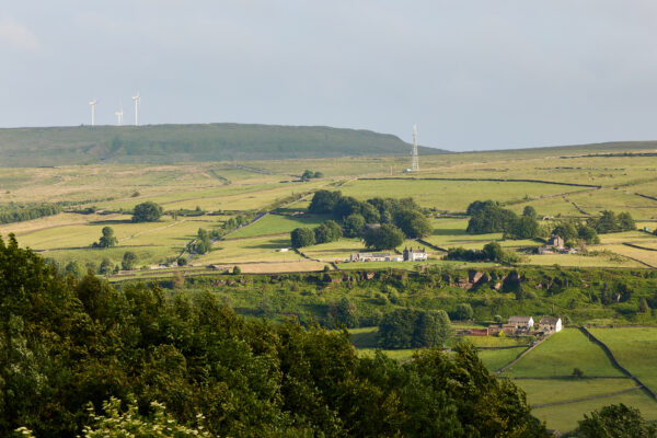 Distant view of rolling green fields divided by stone walls, dotted with farms and trees, with wind turbines and a communications mast visible on the hilltop under hazy sunlight.