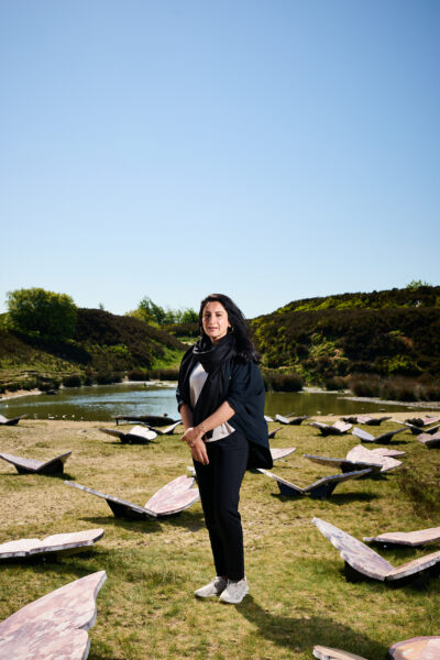 The artist stands among her marble butterfly sculptures near a pond, facing the camera with grassy hills and a clear blue sky in the background.