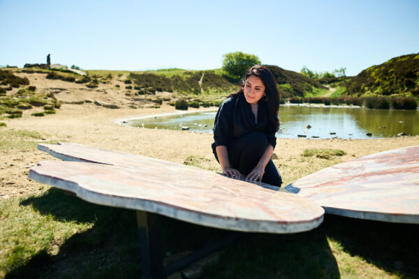 The artist (Meherunnisa Asad) kneels beside one of their large butterfly marble sculptures near a pond, gently touching its patterned surface, with grassy hills and clear skies in the background.
