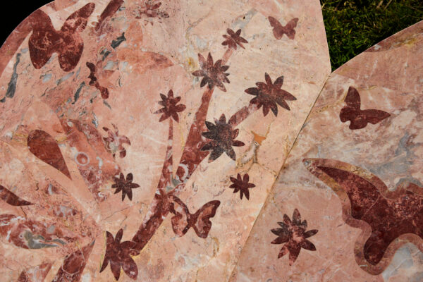 Close-up of engraved pink marble, featuring butterfly and flower motifs in contrasting darker stone, with natural veining and cracks visible across the surface.