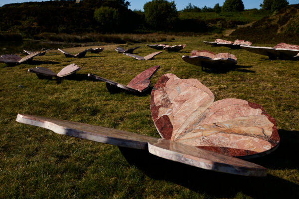Low angle view of pink marble butterfly sculptures spread across a grassy field, with the nearest butterfly’s patterned wings prominently displayed in sunlight against a backdrop of hills and trees.