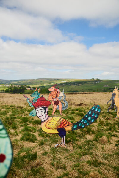 Colourful sculptural figures inspired by medieval paintings of animals, featuring hybrid creatures with human faces and animal bodies, arranged in a grassy landscape with rolling hills under a partly cloudy sky.