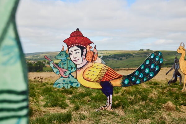 Brightly coloured sculpture of a bird-bodied figure with a human face and red hat, standing in grassy moorland alongside other hybrid creatures under a partly cloudy sky.