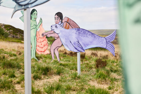 Close-up of outdoor sculptures, featuring a purple seal-like creature mounted on a pole and winged human figures behind, set against grassy moorland and rolling hills.