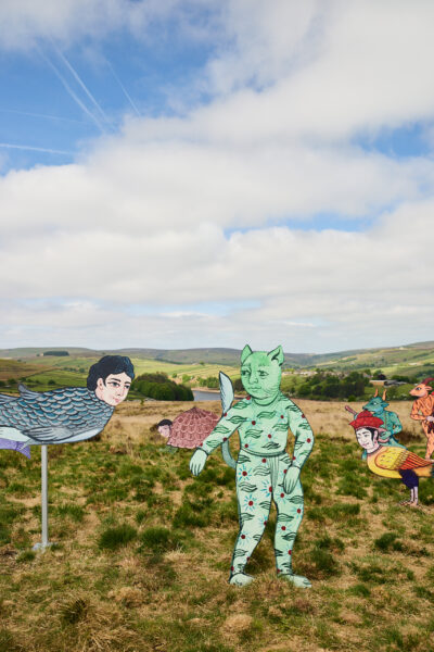 Outdoor installation of brightly coloured sculptures, featuring hybrid creatures such as a green cat-like figure and human-faced fish, arranged on grassy moorland with rolling hills in the background.