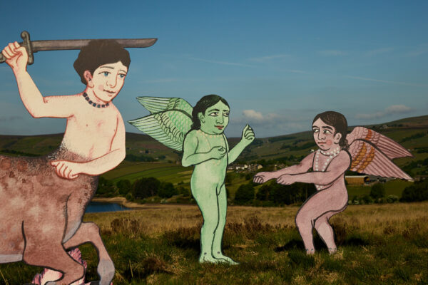 Close-up of three sculptures in a grassy landscape, showing winged humanoid figures and a centaur wielding a sword, styled with bold outlines and painted details against rolling hills.