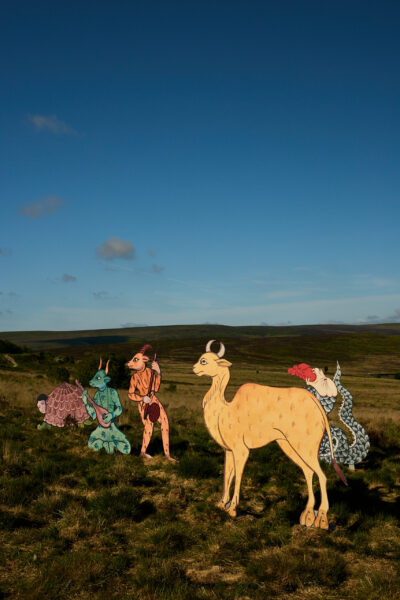 Group of colourful sculptures inspired by medieval animal illustrations, including goat human hybrid horned creatures, horned camel creature and serpent-like figures, displayed in a grassy moorland landscape under a clear blue sky.