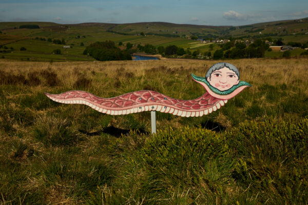 Sculpture of a serpent with the face of an old lady with grey hair and rosy cheeks, painted in red and pink tones with a patterned body, displayed on grassy moorland with hills and a reservoir in the background.