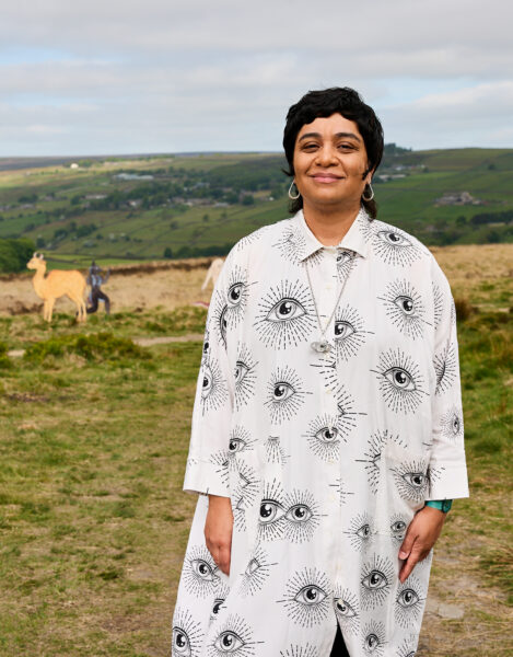 The artist (Monira Al Qadiri) stands smiling outdoors wearing a white coat patterned with black illustrated eyes, with grassy hills and animal sculptures visible in the background.