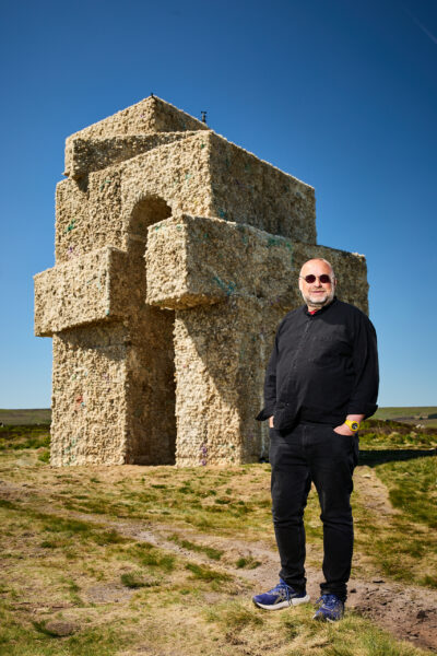The artist (Steve Messam) dressed in black clothing and sunglasses standing in front of large stacked oblong shapes forming a wool arch sculpture on open moorland under a clear blue sky.