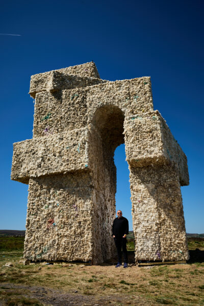 Large stacked oblong shapes forming an arch sculpture made from wool, standing on open moorland under a deep blue sky, with a person in black clothing standing beneath the arch for scale.