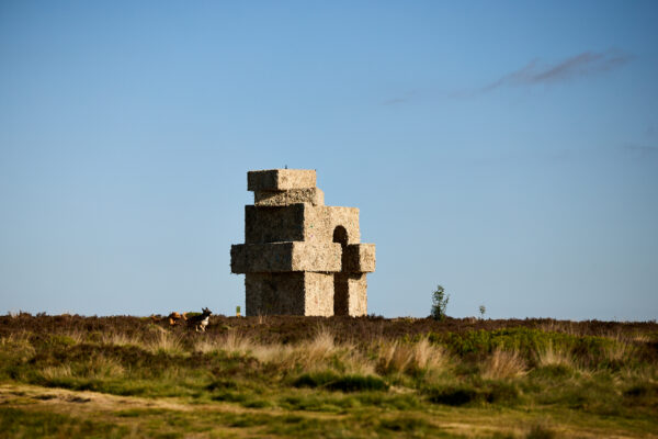 Large stacked oblong shapes, forming an arch sculpture made from wool, positioned on open moorland beneath a clear blue sky, with two dogs running in the foreground.