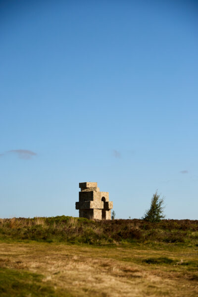 Large stacked oblong shapes forming an arch sculpture made from wool, standing on open moorland with a lone tree nearby, under a clear blue sky.