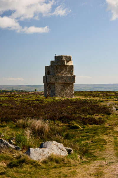 Large stacked oblong shapes, forming an arch sculpture made from wool, standing alone on moorland with heather and grass in the foreground, under a lightly clouded sky with distant hills in view.