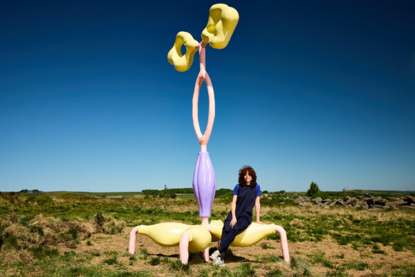 the artist (Vanessa Da Silva) sits on a pastel-coloured abstract sculpture featuring twisting yellow, pink, and purple forms, set against clear blue skies and grassy moorland.