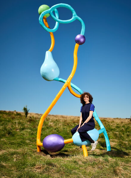the artist (Vanessa Da Silva) sits on a pastel-coloured abstract sculpture featuring twisting yellow, blue, and purple forms, set against clear blue skies and grassy moorland.