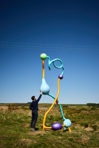 A person reaches up to touch a tall abstract sculpture made of twisting orange, blue and green forms with bulbous shapes, set in grassy moorland under a clear blue sky.