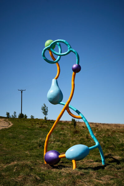 Tall abstract outdoor sculpture composed of bright blue, yellow and orange looping strands, with glossy bulb and sphere shapes in green, purple and light blue, standing on grassy moorland under a clear blue sky.