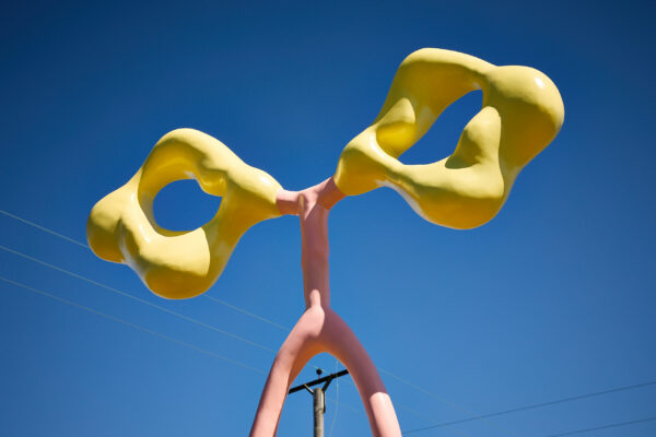 Close-up of glossy abstract outdoor sculpture featuring two bulbous, interconnected yellow forms balanced on a pale pink branching stem, set against a clear blue sky.