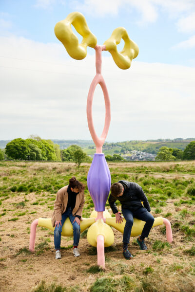 Two people sit and lean forward on the lower yellow parts of a tall pastel abstract sculpture with purple, pink and yellow organic shapes, placed in grassy moorland under a partly cloudy sky.