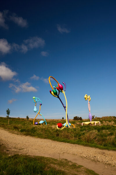 Three tall, brightly coloured abstract sculptures featuring loops, spheres, and organic forms, some resembling flowers stand near a gravel path in open moorland under a clear blue sky with scattered clouds.