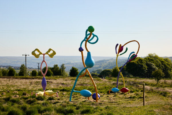 Three tall abstract sculptures in vivid pastel and primary colours, formed from twisting tubes and bulbous shapes, installed on grassy moorland with rolling hills and treetops visible under clear skies.