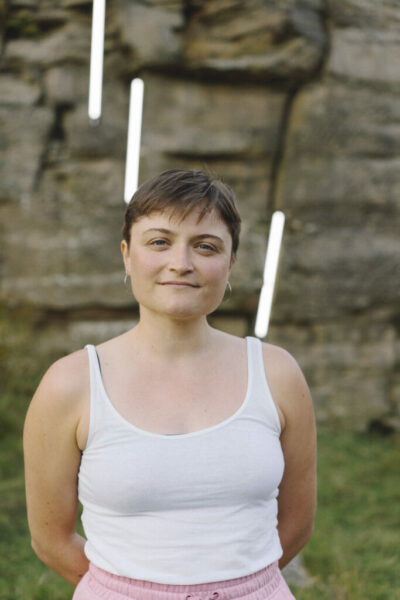 Person with short hair wearing a white tank top and pink pants, standing outdoors against a rocky wall with vertical light tubes positioned behind.