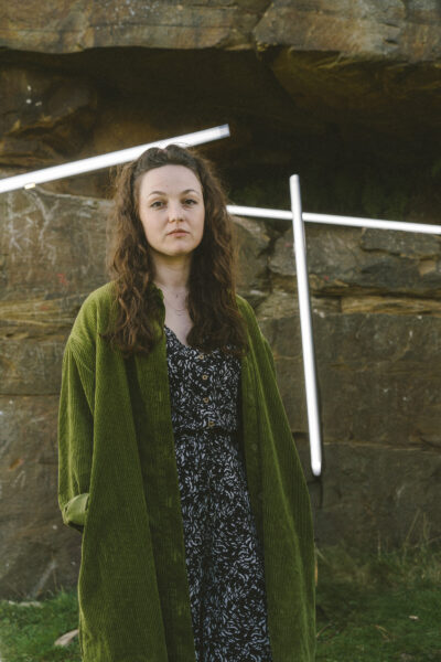 A woman with long curly brown hair wearing a green corduroy jacket over a patterned black dress stands outdoors in front of a rocky wall with diagonal white light tubes behind her.