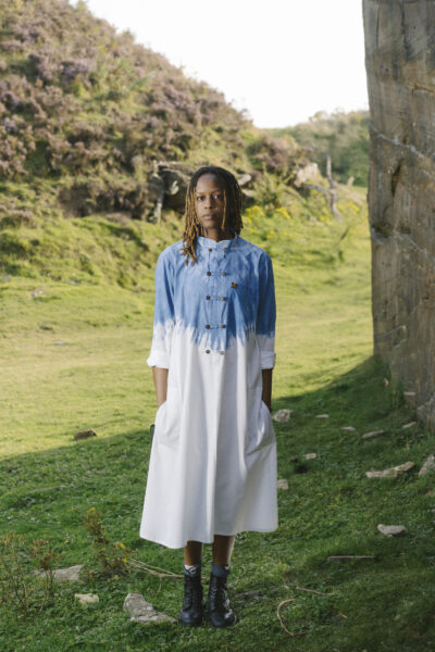 A person standing outdoors on grassy terrain, near a rock face, wearing a white and blue dip-dye style dress with black boots, against a backdrop of hills covered in heather and greenery.